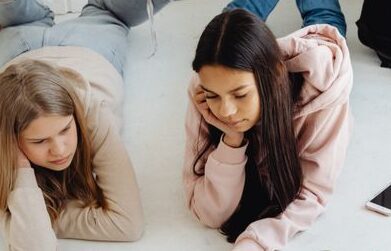 Two teenage girls study together at home using a laptop, surrounded by school supplies.