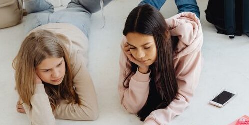 Two teenage girls study together at home using a laptop, surrounded by school supplies.