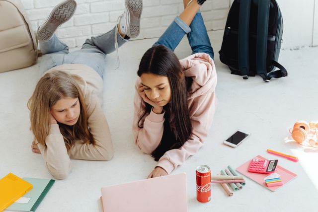 Two teenage girls study together at home using a laptop, surrounded by school supplies.